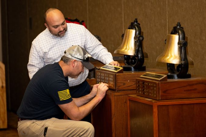 Heartland Division General Manager Tony Fulton watches as Aaron Stich, electrician, applies a nameplate to one of their bells. Heartland Division General Manager Tony Fulton watches as Aaron Stich, electrician, applies a nameplate to one of their bells.
